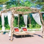 Charming freestanding pergola made from natural wooden vines, draped with white curtains, providing shade over lounge chairs with colorful cushions, set in a lush garden setting