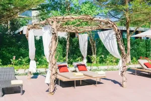 Charming freestanding pergola made from natural wooden vines, draped with white curtains, providing shade over lounge chairs with colorful cushions, set in a lush garden setting