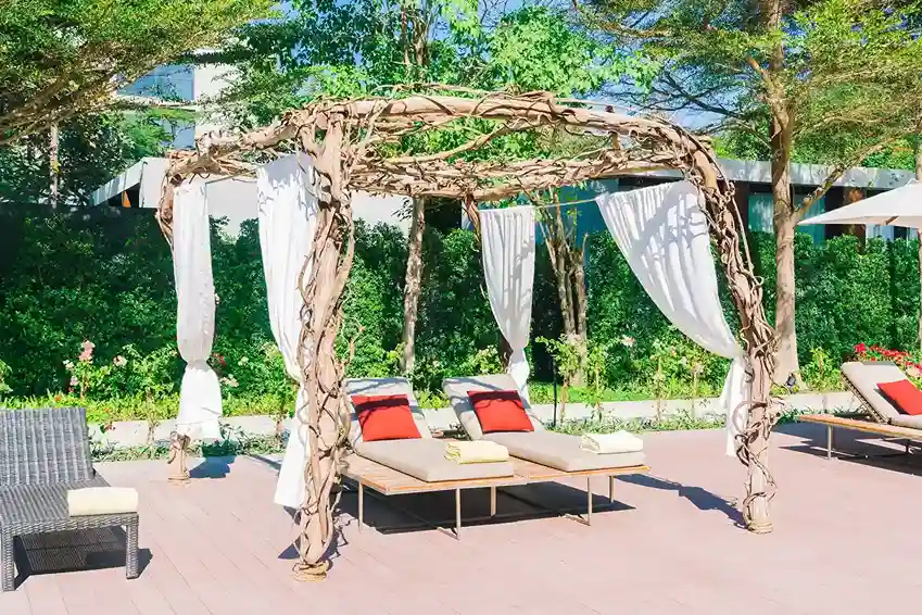Charming freestanding pergola made from natural wooden vines, draped with white curtains, providing shade over lounge chairs with colorful cushions, set in a lush garden setting