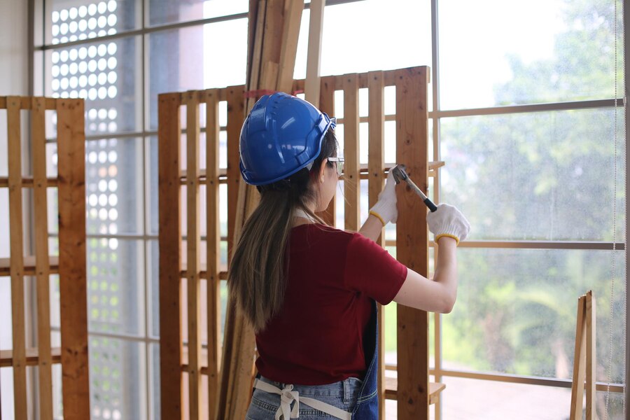 Female carpenter in blue hard hat using a hammer to build a wooden frame by a window, enhancing natural light in the workspace. Ideal for DIY woodworking, home improvement, and interior design projects.