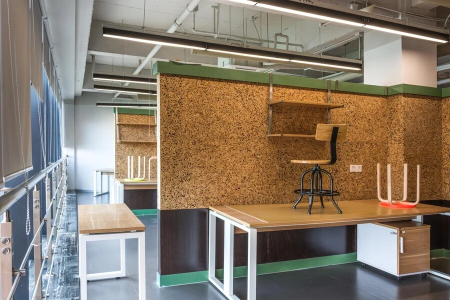 Modern office interior featuring innovative joinery design, with custom-built workstations, shelving, and cork board paneling for added texture. The workspace includes functional elements such as sturdy desks with white metal frames, mounted storage cabinets, and accent lighting. This layout combines practicality with a unique aesthetic, enhancing the space for a collaborative work environment.