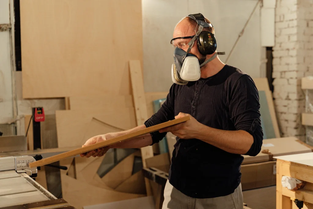 A woodworker in a workshop wearing safety gear, including a respirator mask, ear protection, and goggles, while carefully handling a wooden plank near a table saw. The setting highlights the physical and focused nature of joinery work.