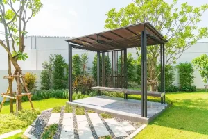 Modern minimalist pergola with a sleek black frame and wooden slat roof, featuring a built-in bench, set in a well-manicured garden with stepping stones and lush greenery.