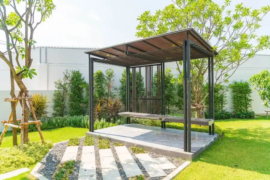 Modern minimalist pergola with a sleek black frame and wooden slat roof, featuring a built-in bench, set in a well-manicured garden with stepping stones and lush greenery.