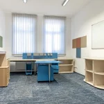 Bright and modern office space with custom joinery, featuring blue and beige furniture tailored for functionality and style. The room includes a spacious desk with integrated storage, open shelving units, and a glass cabinet, all designed to maximize organization. Large windows with blinds allow natural light, creating an inviting work environment ideal for productivity in educational or professional settings.