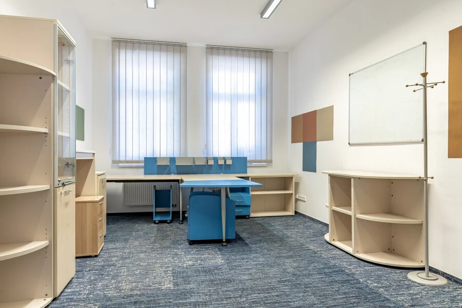 Bright and modern office space with custom joinery, featuring blue and beige furniture tailored for functionality and style. The room includes a spacious desk with integrated storage, open shelving units, and a glass cabinet, all designed to maximize organization. Large windows with blinds allow natural light, creating an inviting work environment ideal for productivity in educational or professional settings.