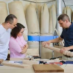 Craftsman shows carved post to couple in workshop. Tools and wood samples visible.