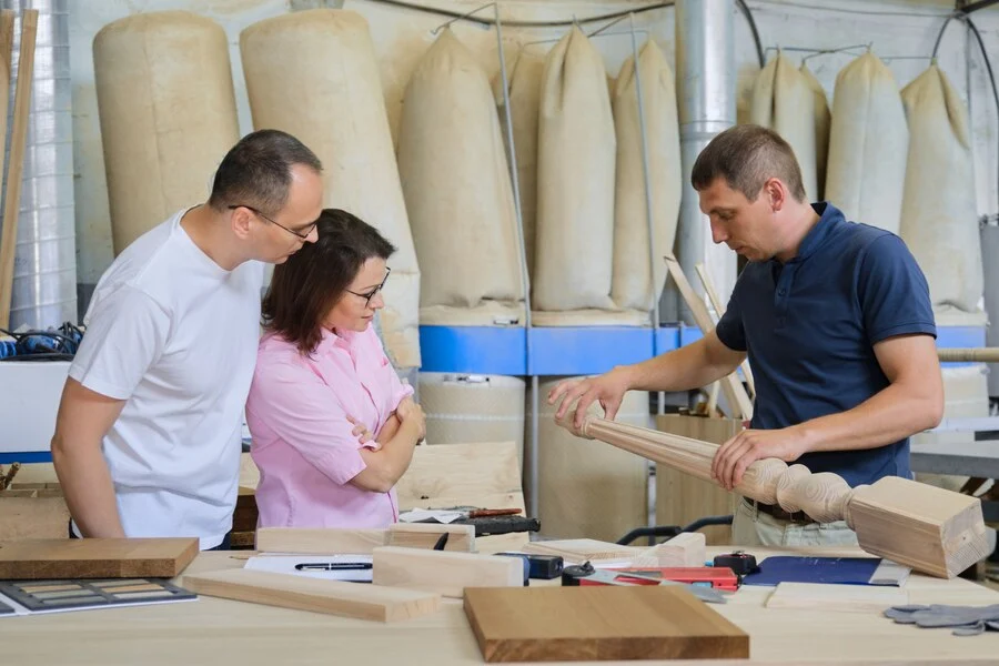 Craftsman shows carved post to couple in workshop. Tools and wood samples visible.