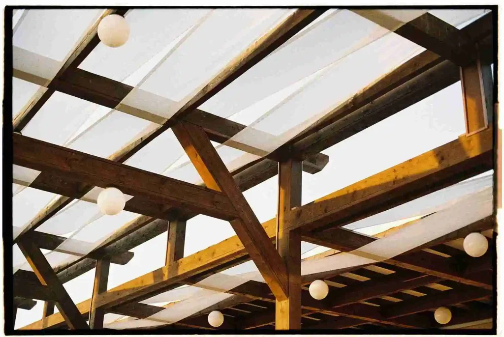 Close-up view of a timber pergola with a transparent roof and hanging round lights, combining rustic charm and modern functionality for outdoor spaces.