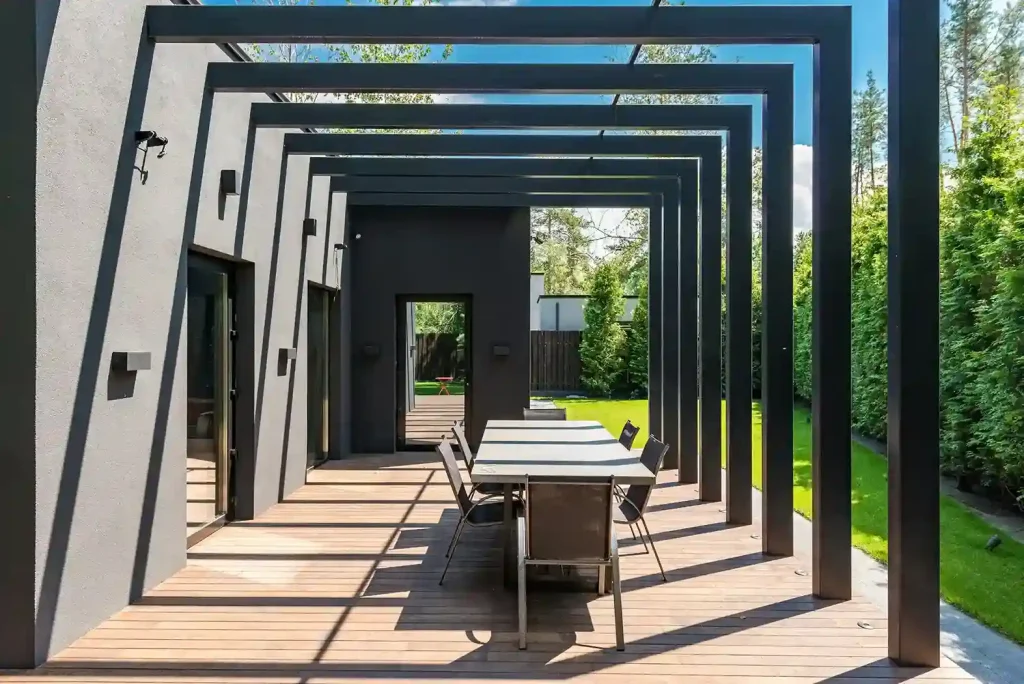 Modern outdoor pergola with sleek black beams creating a stylish framework over a wooden deck, featuring a dining area and surrounded by lush green landscaping, perfect for outdoor living.