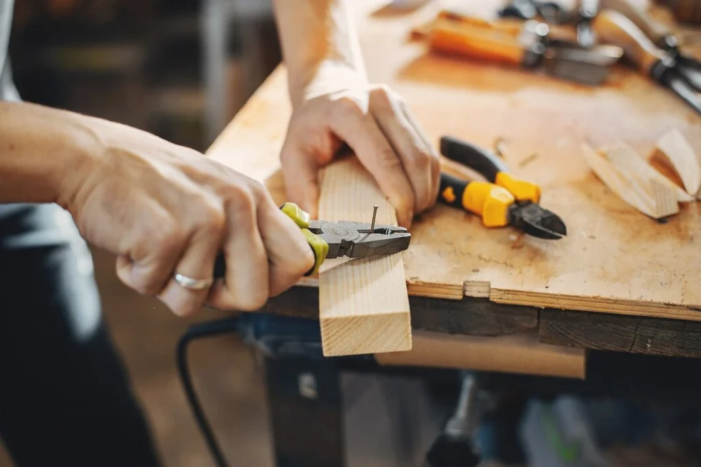 Close-up of a craftsman's hands using pliers to remove a nail from a wooden piece on a workbench, with various tools scattered in the background.