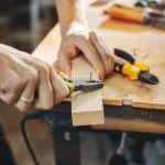 Close-up of a craftsman's hands using pliers to remove a nail from a wooden piece on a workbench, with various tools scattered in the background.