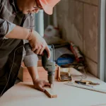 A carpenter wearing a safety helmet using a power drill on a wooden piece in a workshop with tools on the table and natural light coming through a window