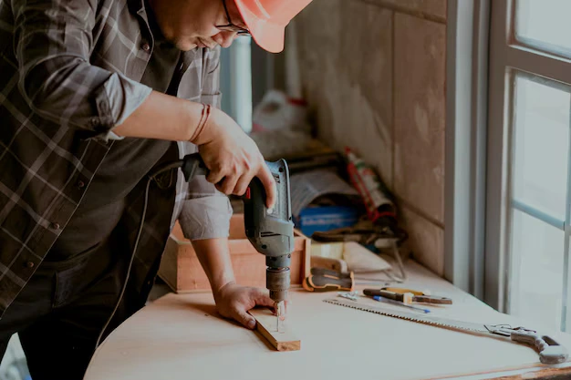 A carpenter wearing a safety helmet using a power drill on a wooden piece in a workshop with tools on the table and natural light coming through a window