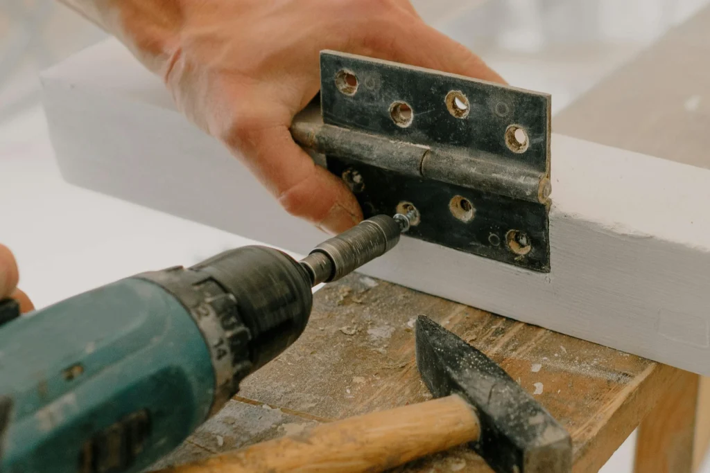 Close-up of a hand using a power drill to attach a hinge to a painted wooden frame, with a hammer resting nearby on a workbench. The image highlights tools and techniques associated with joinery work.