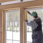 A construction worker using a level to install a large window frame in a house under construction. The scene shows a wintery outdoor landscape through the windows, emphasizing the importance of precise installation and insulation in colder climates.