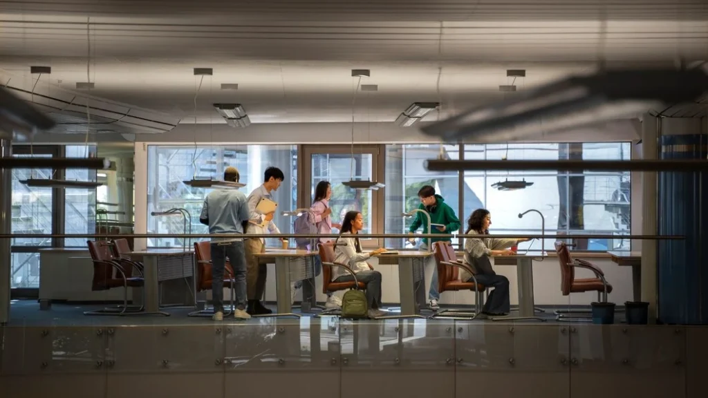 A modern coworking space featuring a group of individuals engaged in various activities. The space includes long desks with built-in lamps and comfortable chairs, set against large windows that let in ample natural light. The environment is open and collaborative, with people working on laptops, reviewing documents, and interacting. The interior design emphasizes functionality and style, creating an inviting atmosphere for productivity and teamwork.