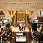 Upscale store: brand displays, escalators, shoppers. Modern, luxury feel.